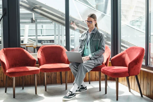 Joven en ropa inteligente se centró en su trabajo sentado en la silla con telón de fondo de vidrio, concepto de coworking - foto de stock