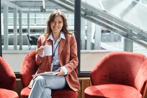 Fröhliche brünette Angestellte sitzt auf Stuhl und trinkt in der Pause ein Getränk, Coworking-Konzept — Stockfoto