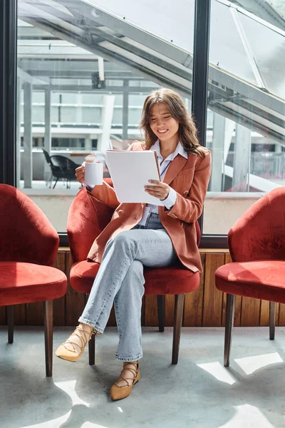 Lächelnde junge Frau im braunen Blazer, die beim Tee ihre Unterlagen überprüft, Coworking — Stockfoto