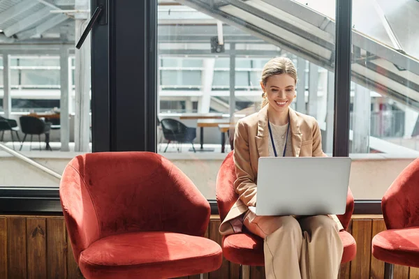 Jovem empregada loira sentada na cadeira trabalhando em seu laptop com fundo de vidro, conceito de co-working — Fotografia de Stock