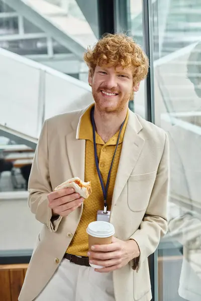 Jeune homme aux cheveux roux joyeux profitant de son café et sandwich à la pause déjeuner, concept de coworking — Photo de stock
