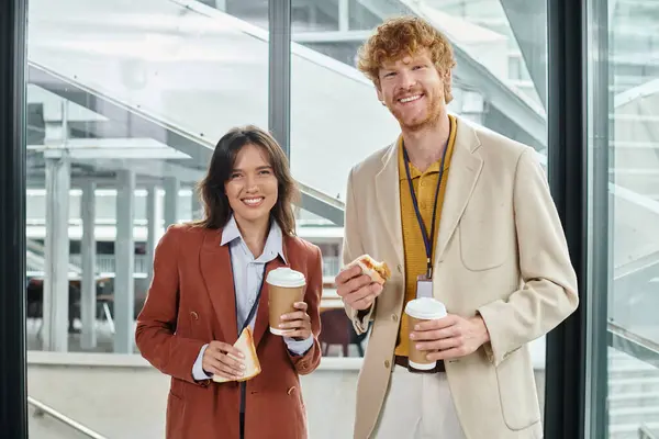 Jovens membros da equipe em trajes inteligentes olhando para a câmera com sanduíches e café, conceito de coworking — Fotografia de Stock