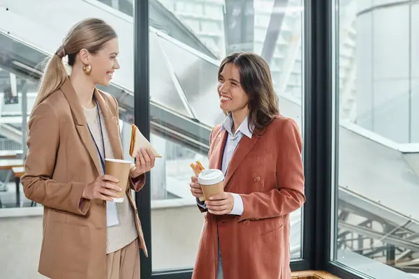 Dos compañeros de trabajo alegres sonriendo el uno al otro y disfrutando de café y sándwiches, coworking - foto de stock