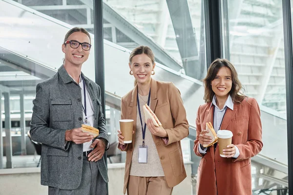 Three colleagues enjoying lunch break with sandwiches and coffee looking at camera, coworking — Stock Photo