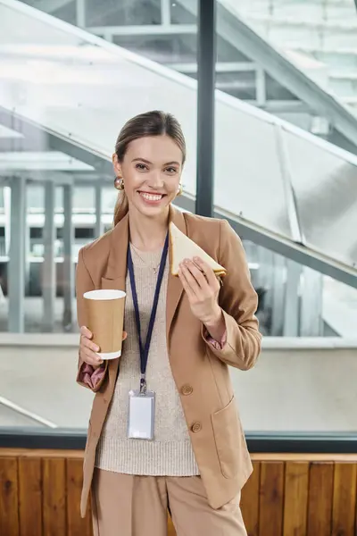 Mujer joven en traje casual inteligente disfrutando de bocadillo y café mirando a la cámara, concepto de coworking - foto de stock