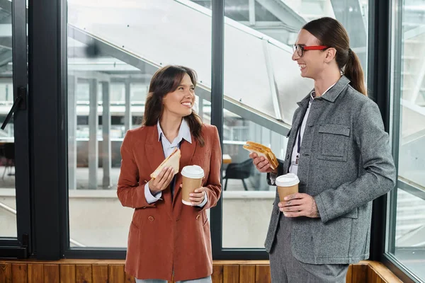 Dois jovens colegas comendo sanduíche com chá no intervalo e sorrindo um para o outro, conceito de coworking — Fotografia de Stock