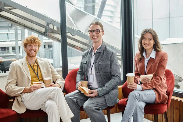 Trois collègues joyeux regardant la caméra et appréciant les sandwichs et le café, concept de coworking — Photo de stock