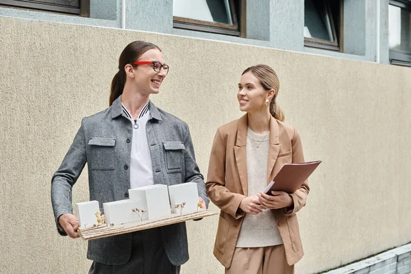 Zwei lächelnde Kollegen, die draußen mit einem maßstabsgetreuen Modell von Gebäude und Papierkram stehen, Designbüro — Stockfoto