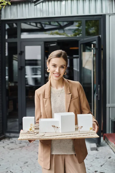 Jolly blonde woman in brown blazer smiling at camera and holding her scale model, design bureau — Stock Photo