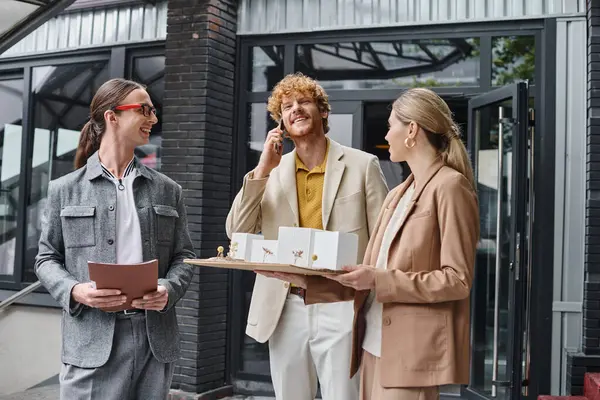 Three young colleagues holding papers and scale model talking on mobile phone, design bureau — Stock Photo