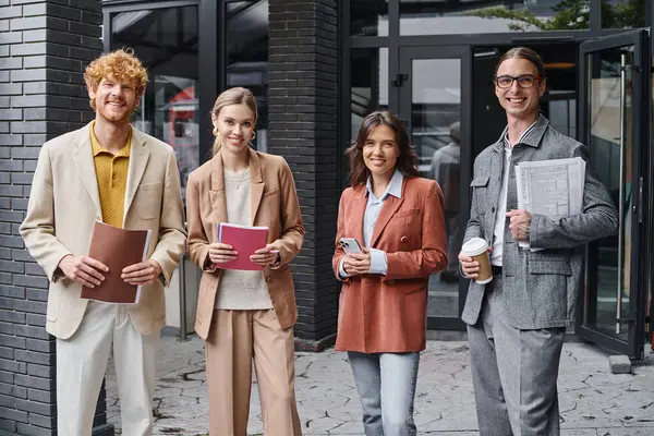 Молодая творческая группа, позирующая снаружи, улыбающаяся перед камерой, держащая бумаги, телефон и кофейную чашку, коворкинг — стоковое фото