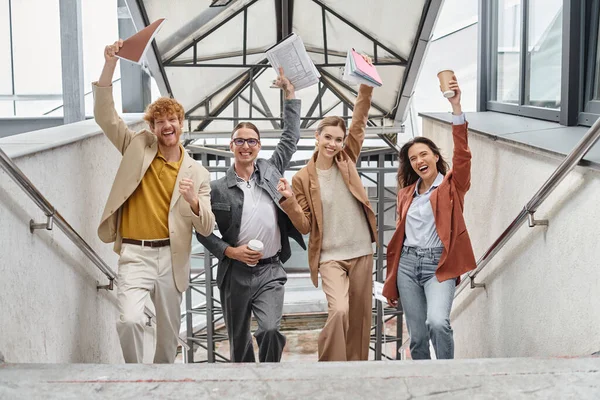Happy cheering teammates posing on staircase with hands with paperwork raised in air, coworking — Stock Photo