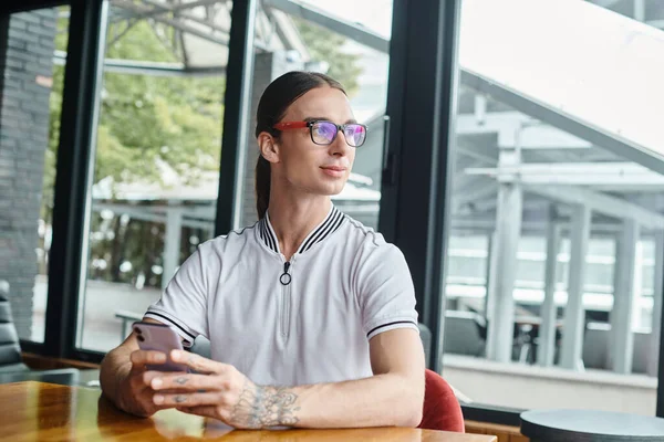 Focused young man in glasses with phone and tattoo looking away with glass background, coworking — Stock Photo