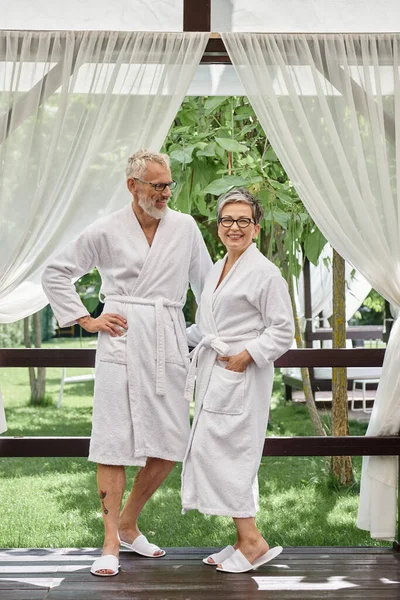 Счастливый мужчина средних лет в очках глядя на жену, зрелая пара, стоящая в белых халатах на курорте — стоковое фото