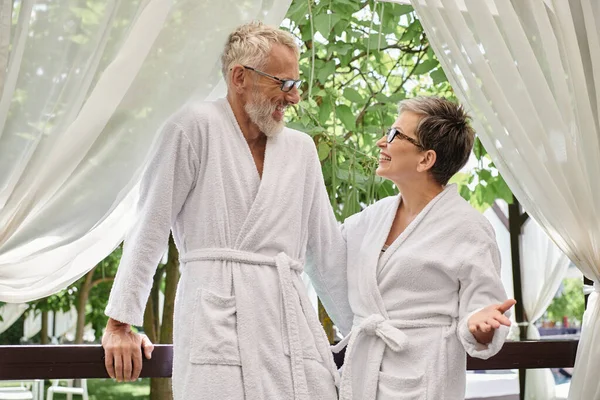 Feliz de mediana edad pareja gafas de pie en túnicas blancas y charlando durante el retiro de bienestar - foto de stock
