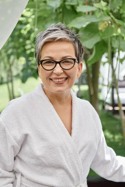 Heureuse femme d'âge moyen en lunettes et robe blanche regardant la caméra pendant la retraite de bien-être — Photo de stock