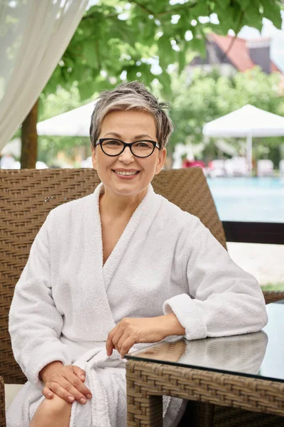 Happy middle aged woman in glasses and white robe sitting at rattan table during wellness retreat — Stock Photo