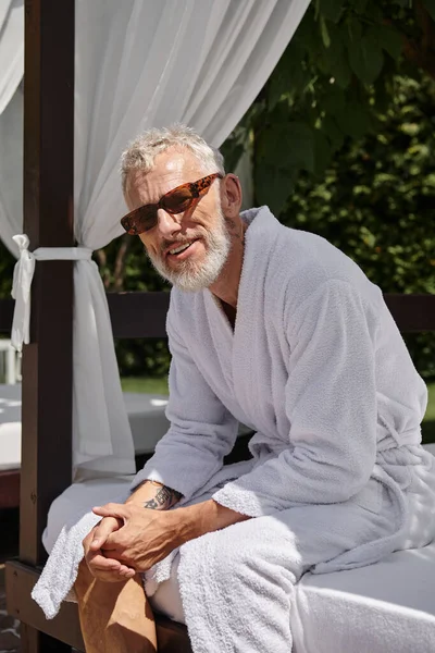 Happy middle aged man in sunglasses and robe resting in private pavilion, wellness retreat in resort — Stock Photo