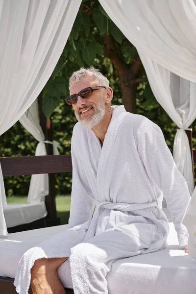 Homme mûr gai en lunettes de soleil et peignoir relaxant dans le pavillon privé, retraite bien-être en station balnéaire — Photo de stock
