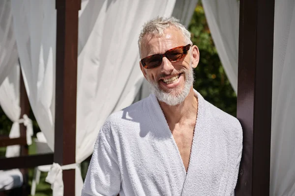Веселий зрілий чоловік в сонцезахисних окулярах і халаті відпочиває в приватному павільйоні, відступає на курорті — стокове фото