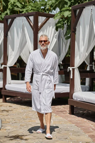 Мужчина средних лет в стильных солнцезащитных очках и белом халате, идущий рядом с частным павильоном в роскошном курорте — стоковое фото