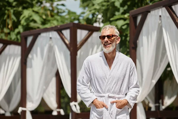 Alegre hombre de mediana edad en gafas de sol y bata posando con las manos en bolsillos en resort de lujo - foto de stock