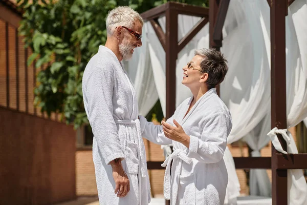 Side view of happy middle aged couple in sunglasses and robes chatting during vacation on resort — Stock Photo
