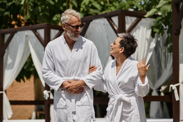 Feliz casal de meia idade em óculos de sol e roupões andando em resort de luxo, conceito de retiro de bem-estar — Fotografia de Stock
