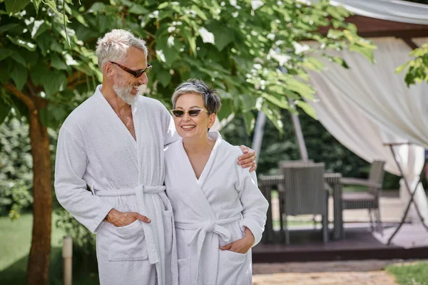 Feliz casal maduro em óculos de sol e roupões de pé em resort de luxo, conceito de retiro de bem-estar — Fotografia de Stock