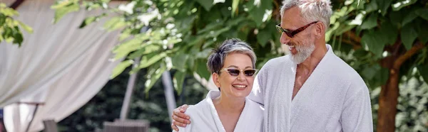 Heureux couple d'âge mûr en lunettes de soleil et peignoirs dans un complexe de luxe, concept de retraite bien-être, bannière — Photo de stock