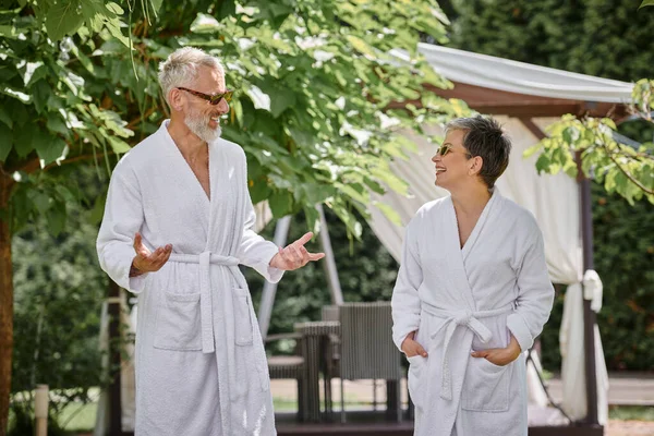 Feliz pareja madura en gafas de sol y túnicas charlando en un resort de lujo, concepto de retiro de bienestar - foto de stock