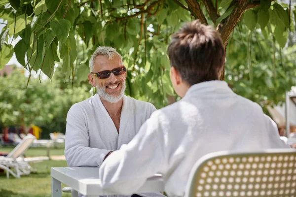 Happy mature man in sunglasses and robe chatting with wife in summer garden, wellness retreat — Stock Photo