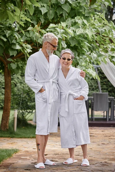 Щасливий зрілий чоловік з татуюванням обіймає дружину в сонцезахисних окулярах і халаті в літньому саду, оздоровчий відступ — стокове фото