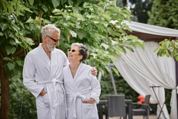 Весёлый зрелый мужчина с татуировкой обнимает жену в солнцезащитных очках и халатах, летний сад, оздоровительный отдых — стоковое фото