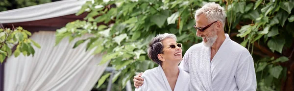Allegro uomo maturo con tatuaggio che abbraccia moglie in occhiali da sole e accappatoio, giardino estivo, banner — Foto stock