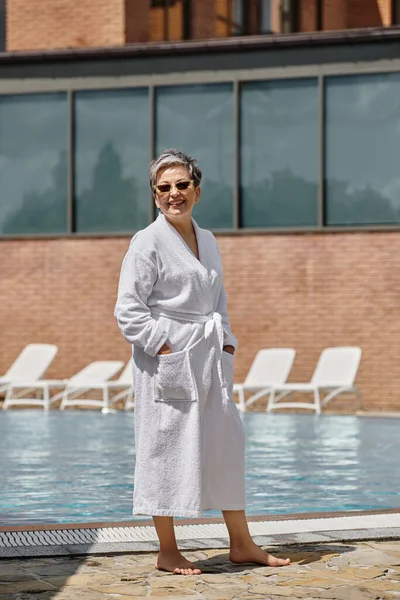 Счастливая женщина средних лет в белом халате и солнцезащитных очках, стоящая у бассейна в роскошном курорте — стоковое фото