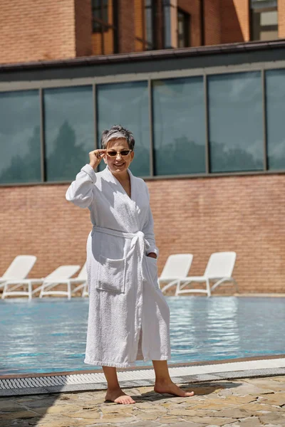 Joyeuse femme d'âge moyen en robe blanche et lunettes de soleil debout au bord de la piscine dans un complexe de luxe — Photo de stock