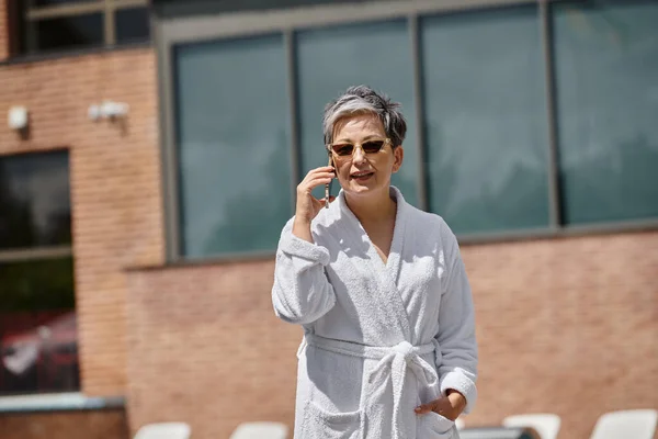 Donna di mezza età in accappatoio bianco e occhiali da sole parlare su smartphone a bordo piscina nel resort di lusso — Foto stock