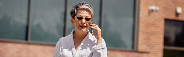 Mujer de mediana edad en bata blanca y gafas de sol hablando en smartphone en resort de lujo, pancarta - foto de stock
