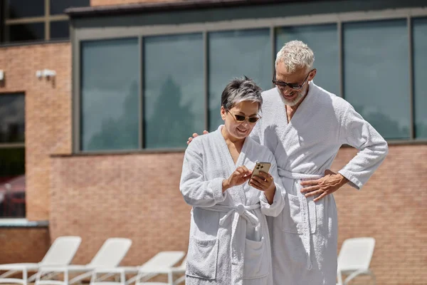Felice donna di mezza età in occhiali da sole utilizzando smartphone vicino al marito a bordo piscina in resort di lusso — Foto stock
