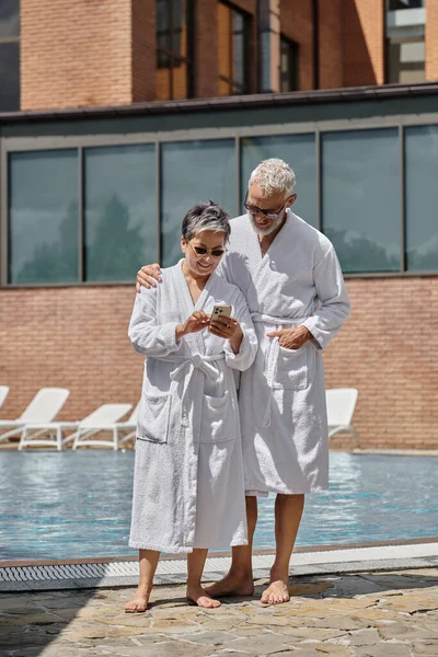 Радость женщина средних лет в солнечных очках с помощью смартфона рядом с мужем у бассейна в роскошном курорте — стоковое фото