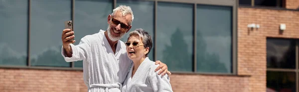 Happy mature couple in robes taking selfie on smartphone in luxury resort, wellness retreat, banner — Stock Photo