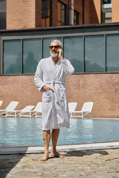 Hombre de mediana edad feliz y tatuado en gafas de sol y bata hablando en el teléfono inteligente en la piscina, retiro - foto de stock