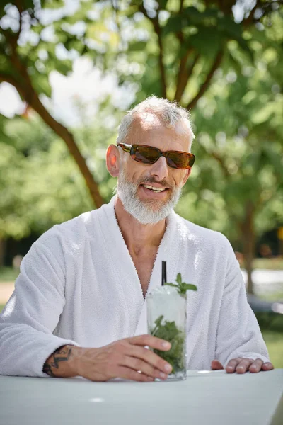 Концепція оздоровчого відступу, веселий зрілий чоловік в сонцезахисних окулярах і халаті насолоджується коктейлем у відпустці — стокове фото