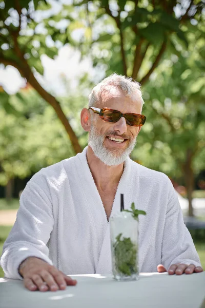Concepto de retiro de bienestar, hombre maduro feliz en gafas de sol y bata disfrutando de cóctel en vacaciones - foto de stock