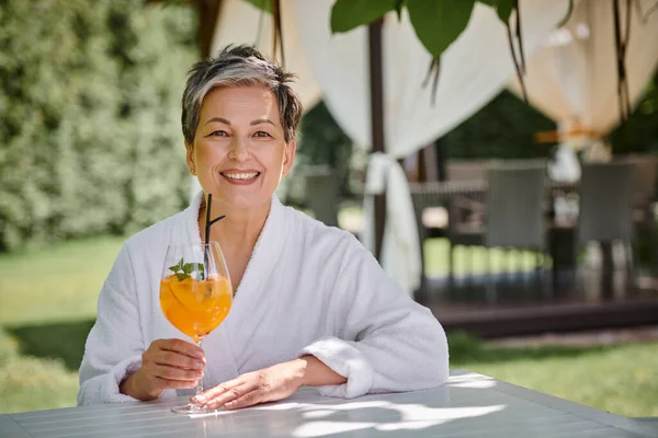 Concept of wellness retreat, happy mature woman in white robe enjoying cocktail on vacation — Stock Photo