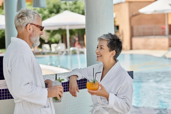 Happy middle aged couple in sunglasses and robes holding cocktails at poolside, wellness retreat — Stock Photo