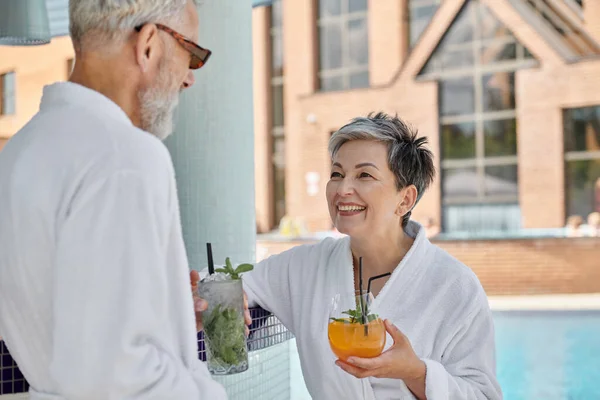 Cheerful middle aged couple in sunglasses and robes holding cocktails at poolside, wellness retreat — Stock Photo