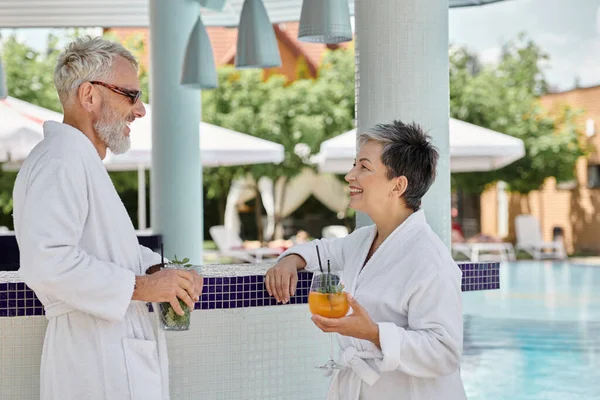 Cheerful mature couple in sunglasses and robes holding cocktails at poolside, wellness retreat — Stock Photo