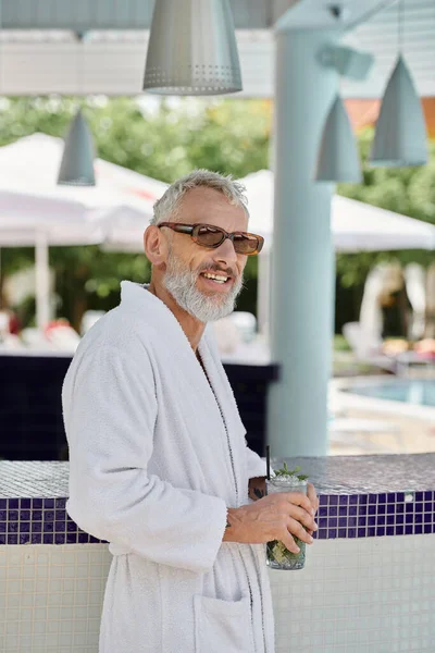 Cheerful mature man in sunglasses and robe holding mojito cocktail at poolside, wellness retreat — Stock Photo